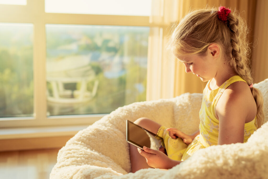 Happy blonde girl reading something on her ipad while seated on a puff